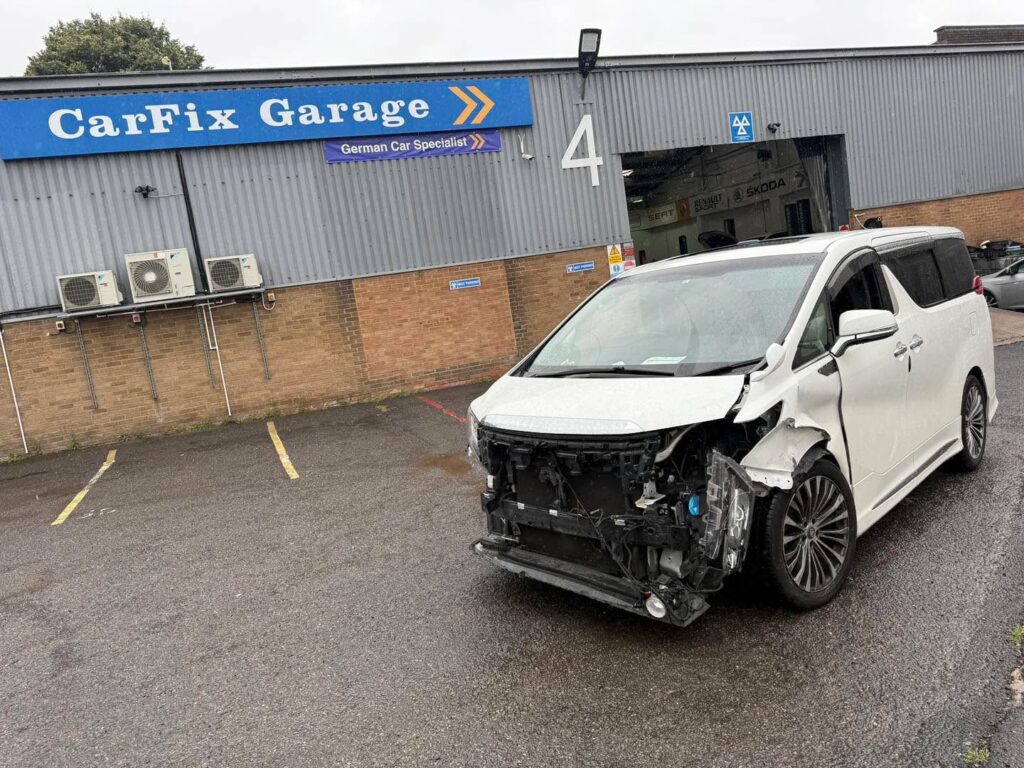 Un minivan alb (ca un Toyota Alphard, des folosit pentru PHV) cu partea frontală avariată, parcat pe o stradă din Londra.